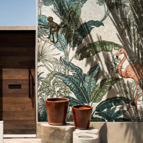 Outdoor jungle mosaic mural with flamingo, monkey and lush tropical leaves beside a wooden gate and clay pots, sunlit in a modern courtyard entrance.