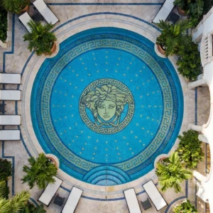Overhead view of luxury round courtyard pool with Medusa mosaic and Greek key border, surrounded by arches, palm plants and sun loungers.