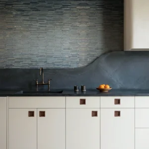 Modern kitchen with charcoal linear tile backsplash and curved dark stone countertop, paired with white cabinets, bronze faucet, and minimal decor.