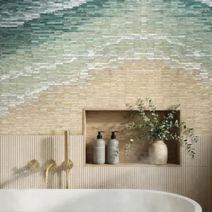 Spa bathroom with gradient mosaic tile wall above freestanding tub, brass fixtures, and wood niche shelf with bottles and greenery.