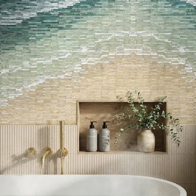 Spa bathroom with gradient mosaic tile wall above freestanding tub, brass fixtures, and wood niche shelf with bottles and greenery.