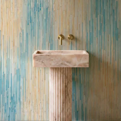 Vertical ombre glass mosaic wall in soft blue and beige tones behind a stone pedestal sink with brass fixtures in a modern bathroom.