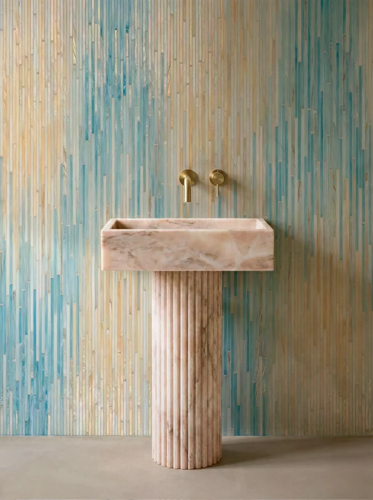 Vertical ombre glass mosaic wall in soft blue and beige tones behind a stone pedestal sink with brass fixtures in a modern bathroom.