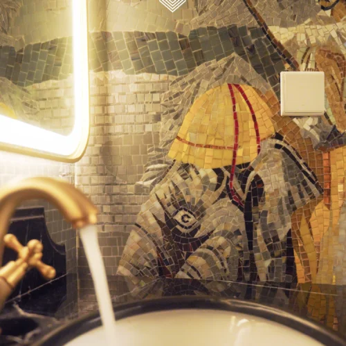 Elephant mosaic mural in warm gold and neutral tones installed behind a bathroom sink with soft lighting.