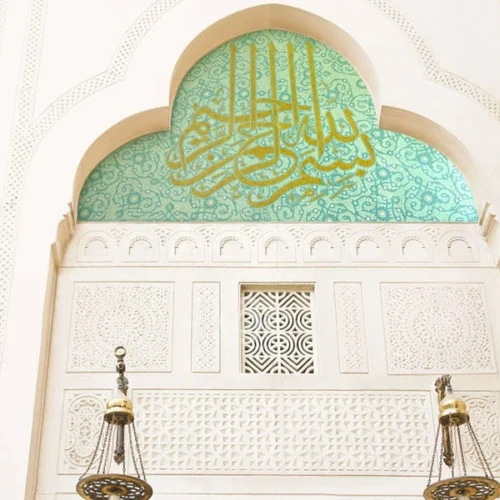 Islamic arch with golden Arabic calligraphy on green mosaic background featuring floral patterns and intricate mosque interior detailing.