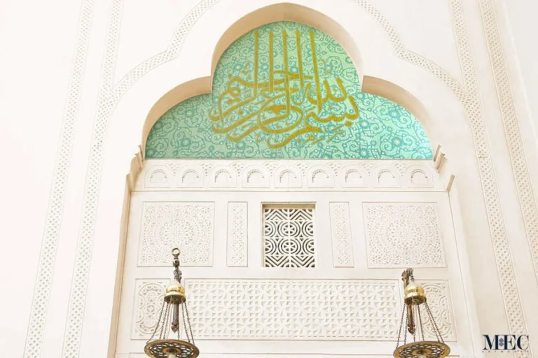 Islamic arch with golden Arabic calligraphy on green mosaic background featuring floral patterns and intricate mosque interior detailing.