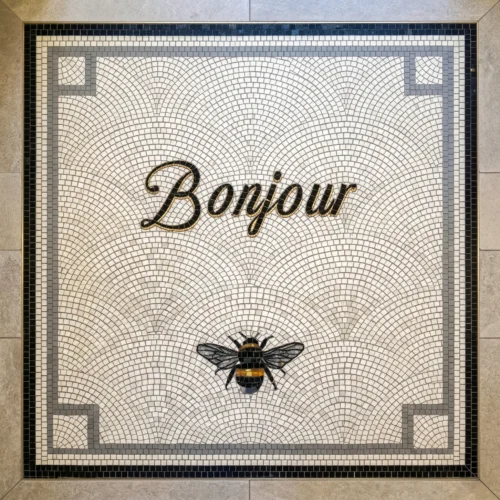 Mosaic tile panel with “Bonjour” script and decorative bee on patterned background.