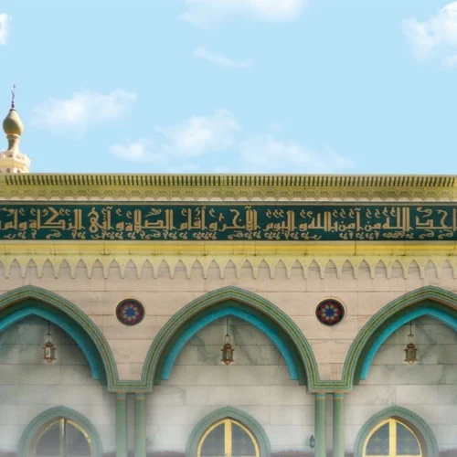 Islamic mosque façade with green mosaic calligraphy band in Arabic script above arched windows, reflecting traditional architectural design.