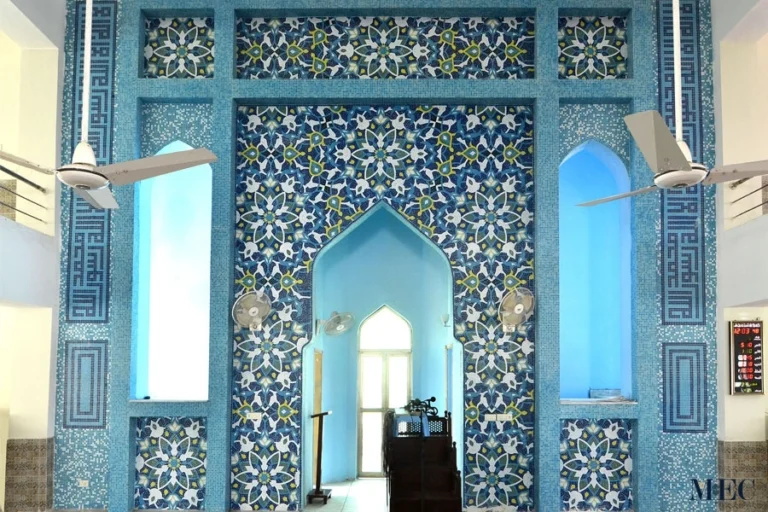 Blue Islamic mosaic wall with floral and geometric patterns surrounding a mosque mihrab, showcasing traditional craftsmanship and symmetrical design.