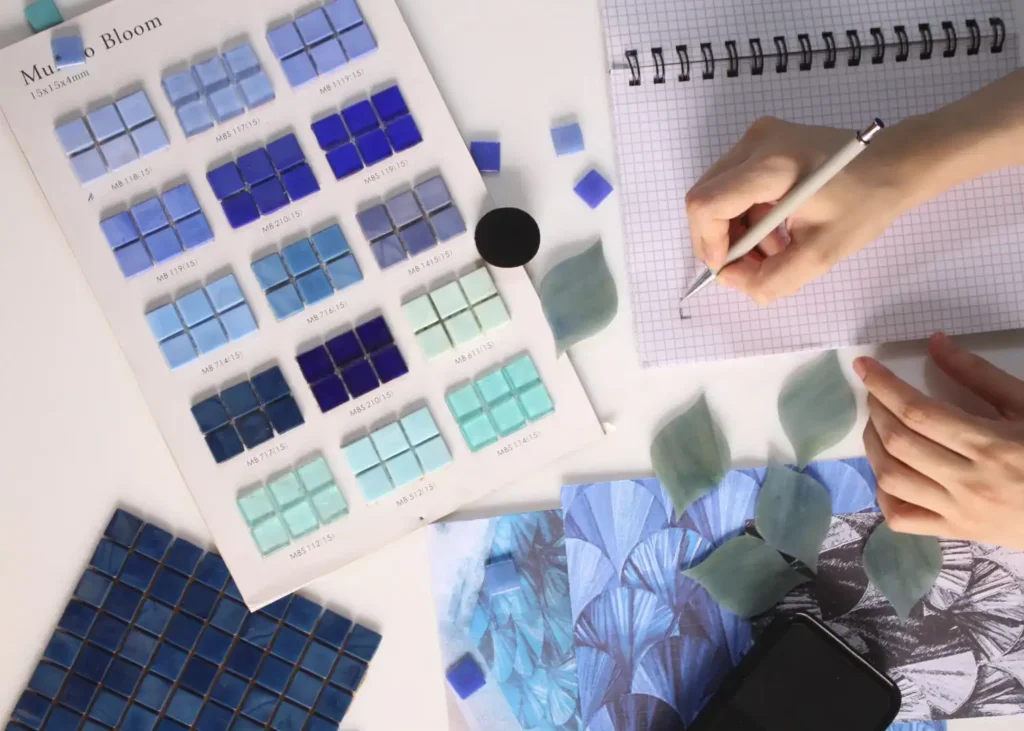 An overhead shot showing a hand working on grid paper along with a color palette carrying different tones of blue