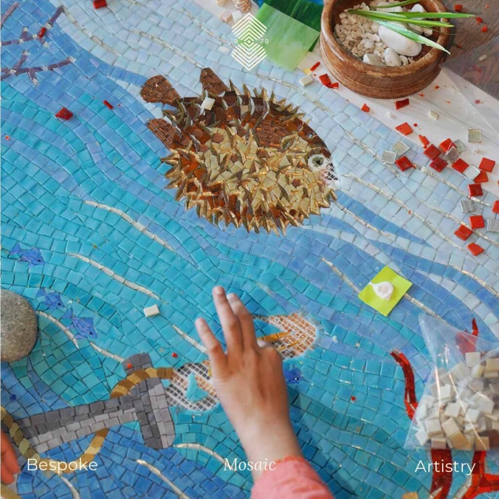 A hand meticulously working on a luxury marine mosaic art with blue glass tiles along with a pufferfish above. 