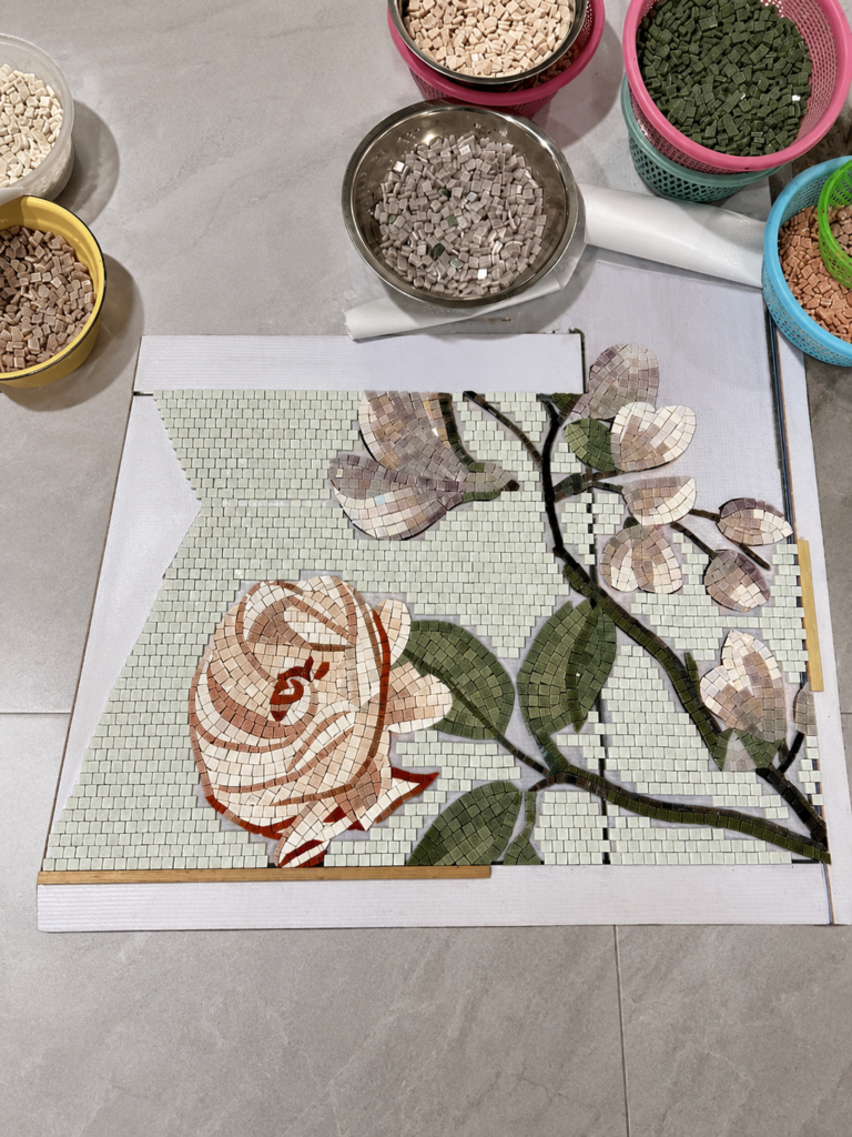 Overhead view of a mosaic artisan assembling small square glass tiles into a pink rose and green leaf design, with small bowls of tile pieces nearby.