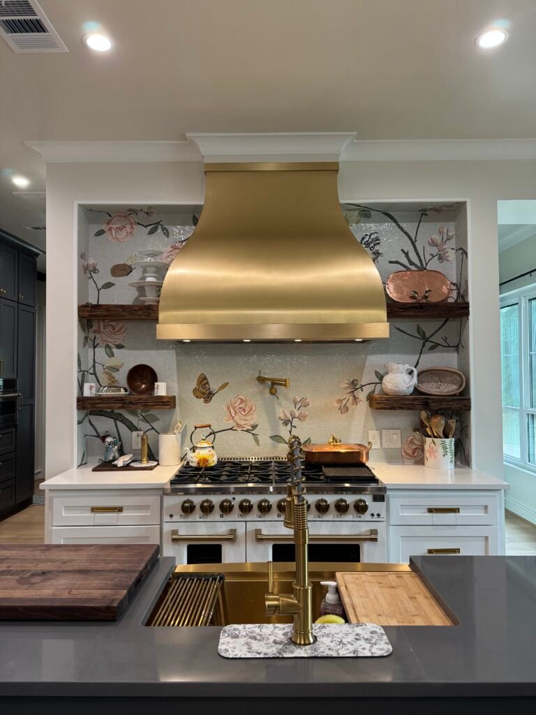 Frontal view of a luxury kitchen featuring a brushed gold range hood, custom glass mosaic backsplash with blush pink roses and butterflies, dark floating wood shelves, and a gold island faucet overlooking the range.