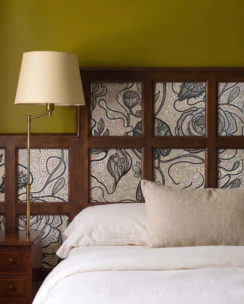 Bed side with beige pillow and white sheets, followed by a cream lamp with a golden handle. Behind it is a grey and dark floral mosaic art against a light background.
