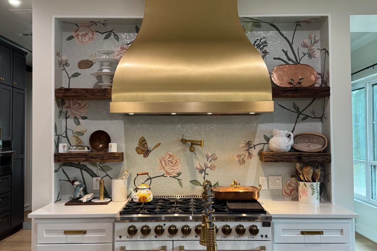 A centered shot of an elegant kitchen viewed from behind the island, showing a brushed gold range hood, handcrafted floral glass tile backsplash, rustic dark wood floating shelves, and a matching gold faucet.