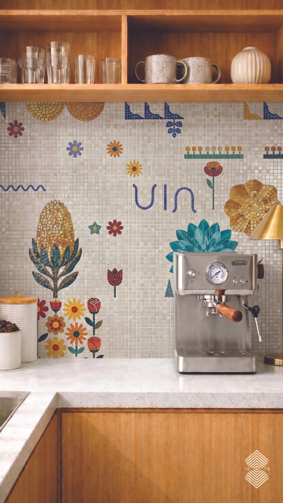 A modern kitchen counter with wooden base and white marble. A coffee station vibe with espresso machine, showing a playful, vibrant mosaic tile backsplash on a cream grid background, decorated with whimsical floral motifs in bright yellow, green, red, and blue.