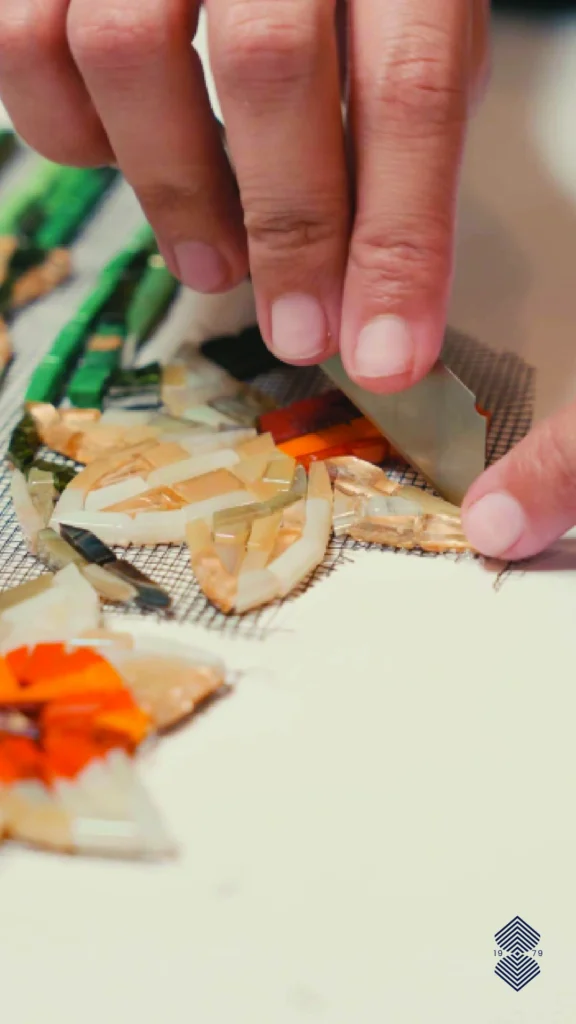 A close-up of a person's hands using a small tool to carefully place and adjust tiny, uniquely cut pieces of glass tile, actively creating a delicate floral mosaic design.
