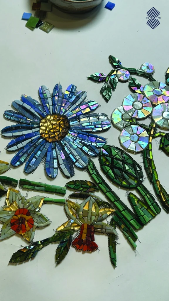 A crystal clear close up of a manually hand-cut floral mosaic artwork in progress, showcasing stunning and luxurious glass mosaic pieces in blue, yellow, green, and red.