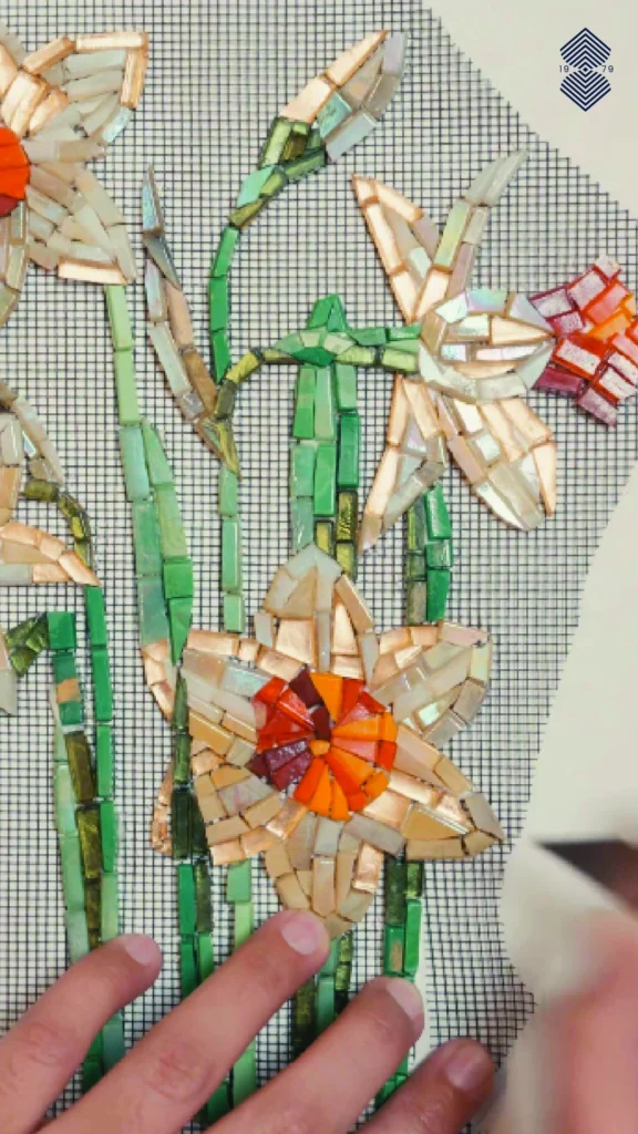 A hand working on a yellow hand-cut floral mosaic piece, with green stems.