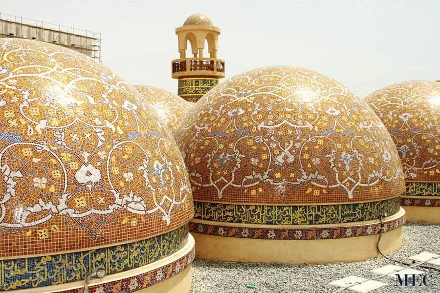 Multiple domes covered in intricate gold and white floral mosaics, featuring a band of gold Arabic calligraphy beneath. 