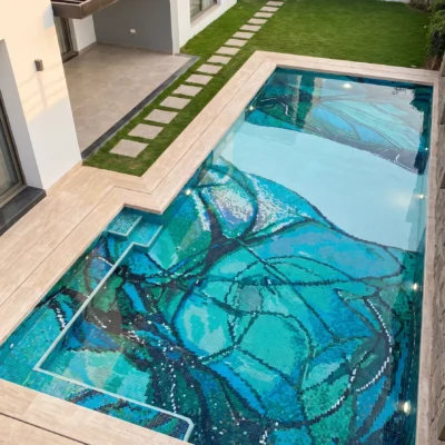 Luxury swimming pool featuring a handcrafted glass mosaic with large botanical leaf patterns in turquoise, aqua, emerald, and deep blue tones.