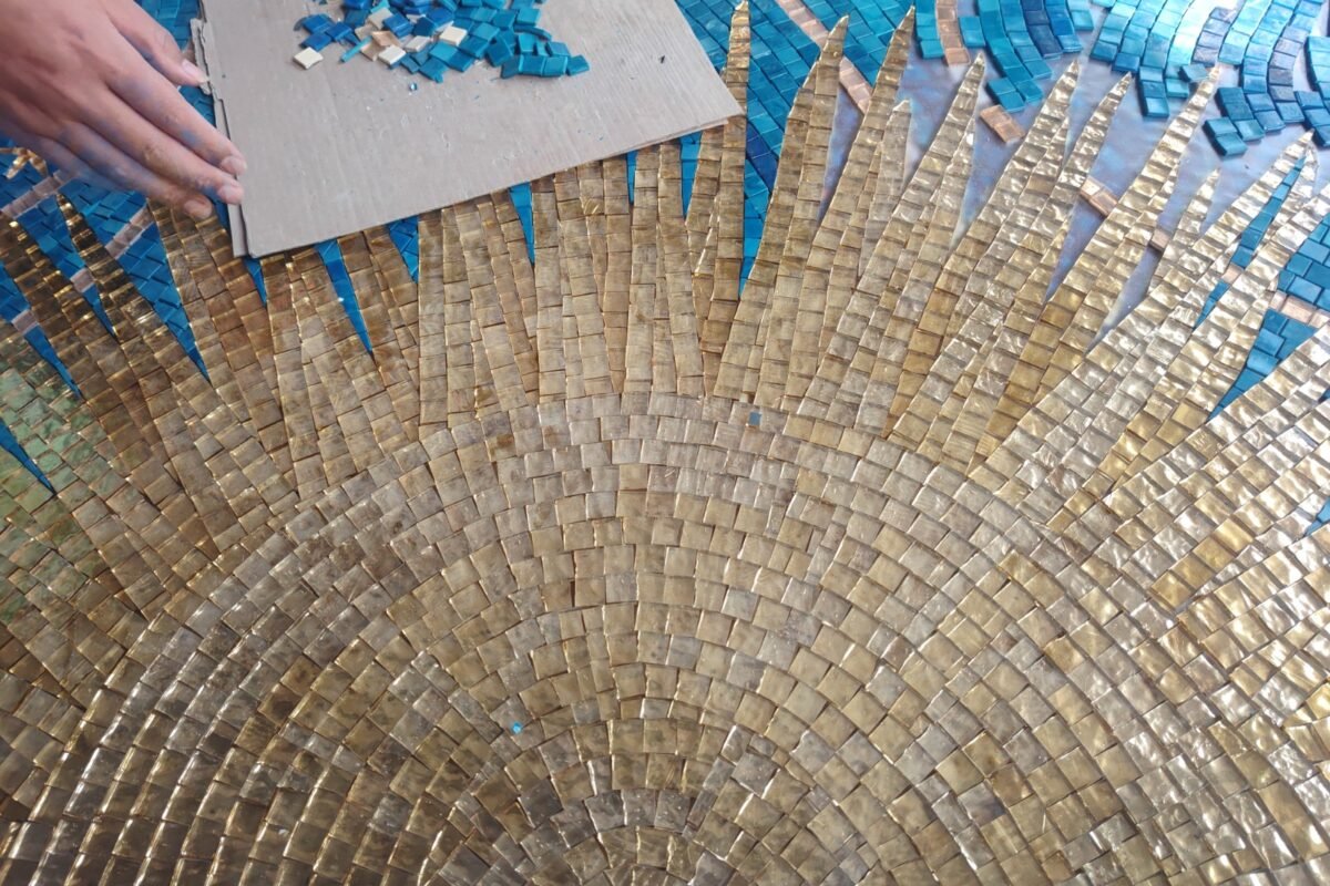 Someone's hands placing small blue tiles next to the bright gold rays of a mosaic sun.