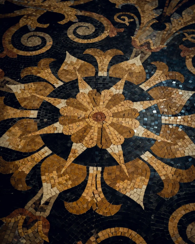 A close-up of a completed mosaic rug featuring a large, intricate golden-brown floral starburst design against a dark black and blue background with swirling motifs.