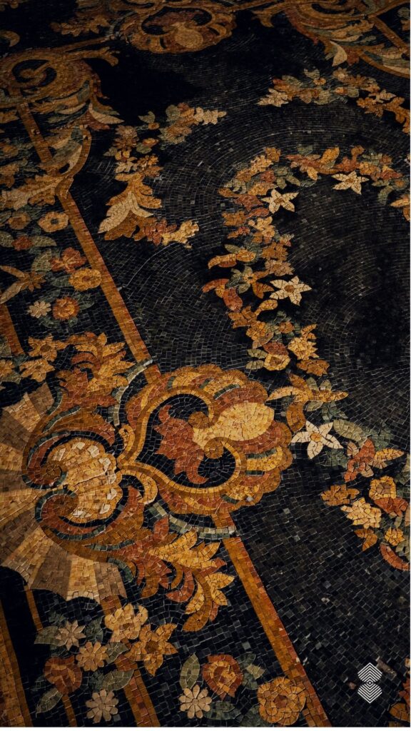 A detailed top-down view of a dark marble mosaic floor featuring intricate floral scrollwork and border patterns in shades of gold, amber, and earth tones.
