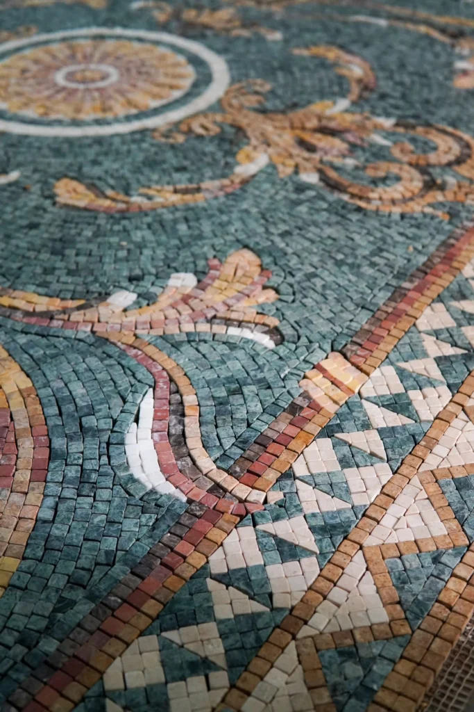 A detailed close-up of a mosaic surface showing a teal blue background with swirling beige and pink leafy patterns, bordered by a geometric design of small triangles.