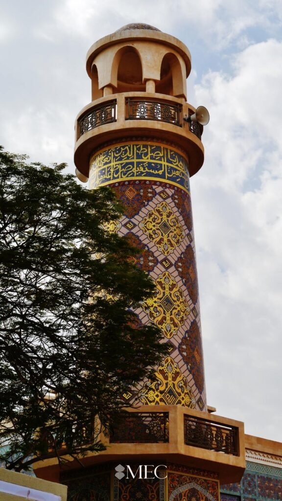 A tall minaret tower decorated with intricate gold, brown, and blue mosaic patterns followed by a band of Arabic calligraphy.