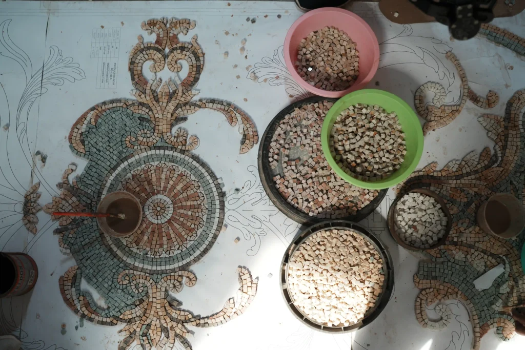 An overhead shot of a large-scale mosaic artwork in progress on a printed template. The intricate design is partially filled with teal, beige, and pink stones. Several brightly colored bowls containing loose, sorted mosaic tiles and a cup with a brush are scattered across the workspace.