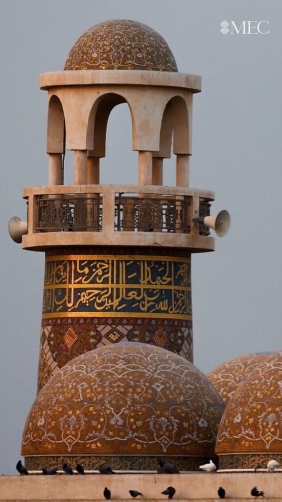 A minaret featuring gold Arabic calligraphy and loudspeakers, rising above domes covered in intricate gold and white floral mosaics.