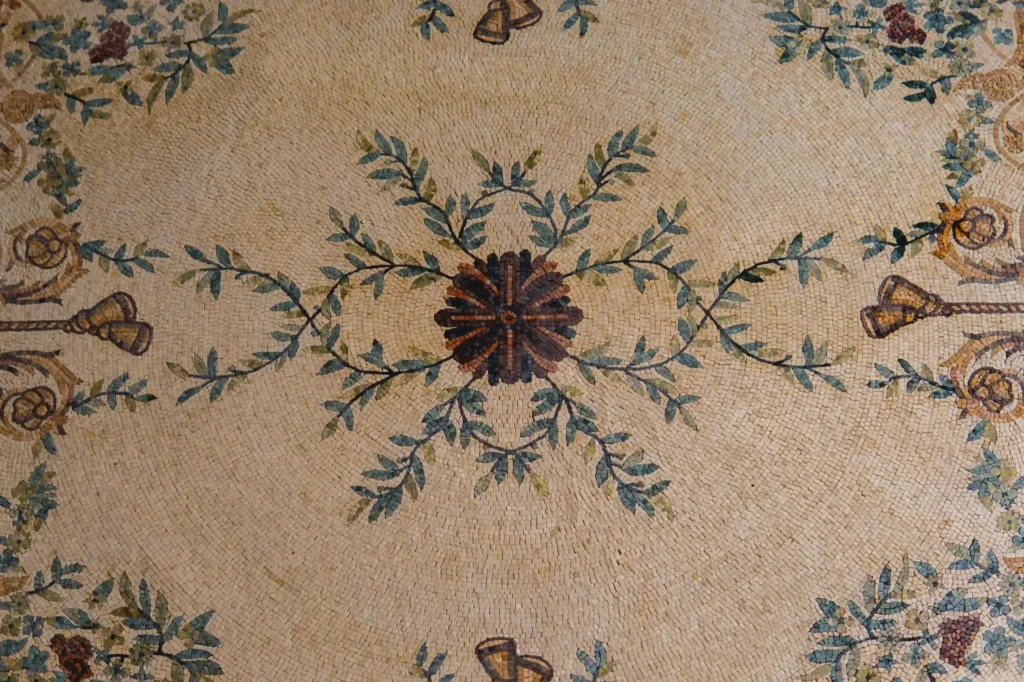 A detailed view of a cream-colored stone mosaic featuring a central dark floral medallion with symmetrical green leafy vines.
