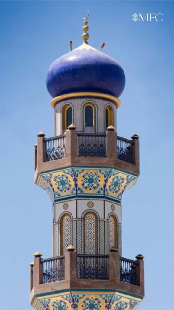 A tall minaret with intricate blue, yellow, and white mosaic patterns, topped by a solid blue dome and a golden crescent.