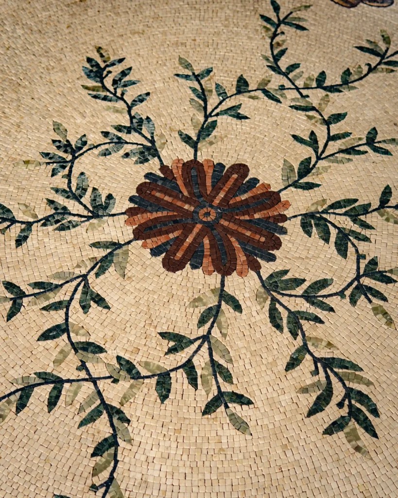 A completed mosaic featuring a light beige background with a central brown, multi-petaled flower design. Green leafy vines branch outwards from the center in a symmetrical pattern.