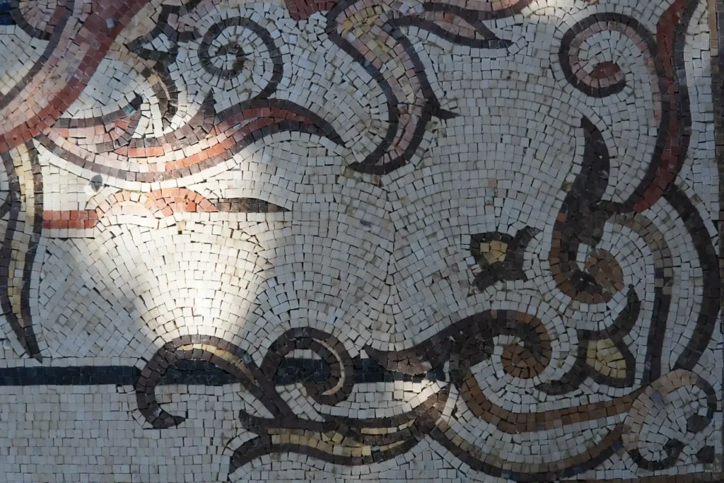 A close-up of a light cream mosaic artwork displaying swirling, acanthus leaf-style patterns in dark brown and terracotta stone tiles, with sunlight hitting the left side.