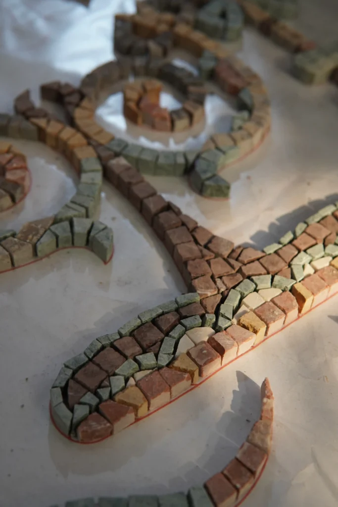 A macro shot of small, square mosaic stones in earthy brown, green, and beige tones, glued in curved lines following a red outline on a flat surface.