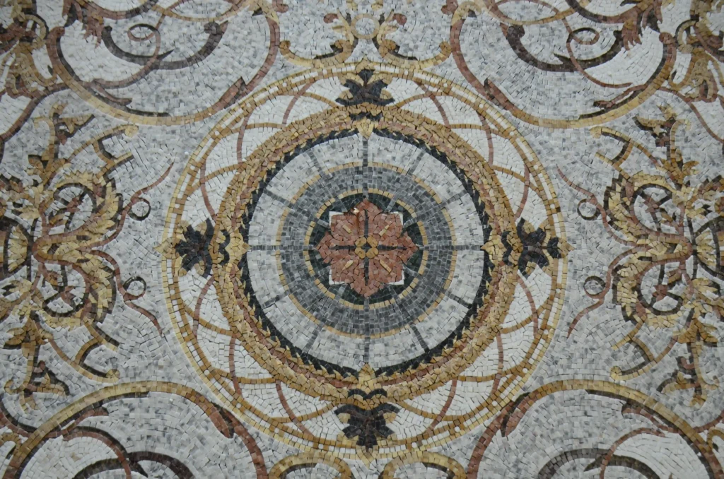 A wide view of a highly detailed mosaic medallion featuring concentric circles in grey, white, and brown, with a small central flower and ornate, symmetrical swirling patterns around the outer edges.
