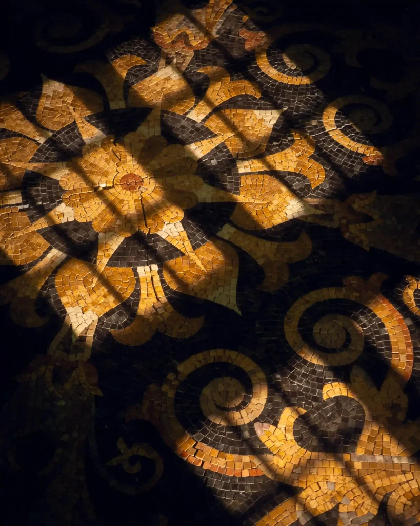 Dramatic shafts of sunlight stream across a dark mosaic floor, illuminating a large, central star-shaped floral medallion made of golden-yellow and amber stone tiles.
