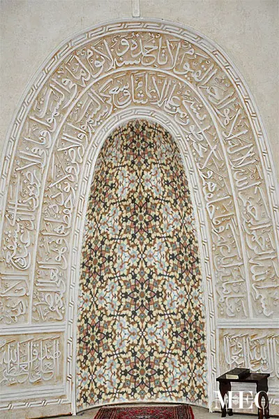 A recessed arched wall niche (mehrab) framed by heavily carved Arabic calligraphy, featuring an intricate geometric and floral mosaic interior.