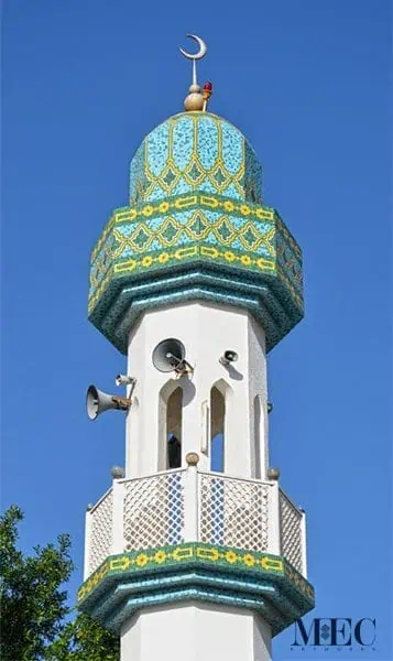 A white minaret tower featuring loudspeakers and balconies accented with blue, green, and yellow mosaics.