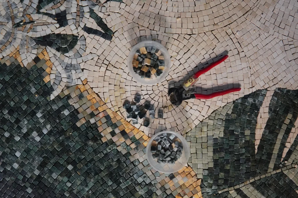 A top-down view of a partially completed mosaic artwork featuring green leafy sections and beige backgrounds. A red-handled tile nipper tool and two small plastic cups filled with loose grey and beige stones sit on the unfinished area.