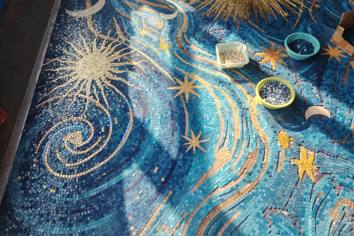 A large starry night mosaic in blue and gold tones spread across a sunlit floor, with three small baskets of loose tiles resting on its surface.