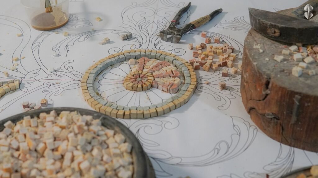 A close-up view of a partially completed circular stone mosaic resting on a hand-drawn paper template. Pliers, loose square stone tesserae, and a wooden cutting block sit nearby.