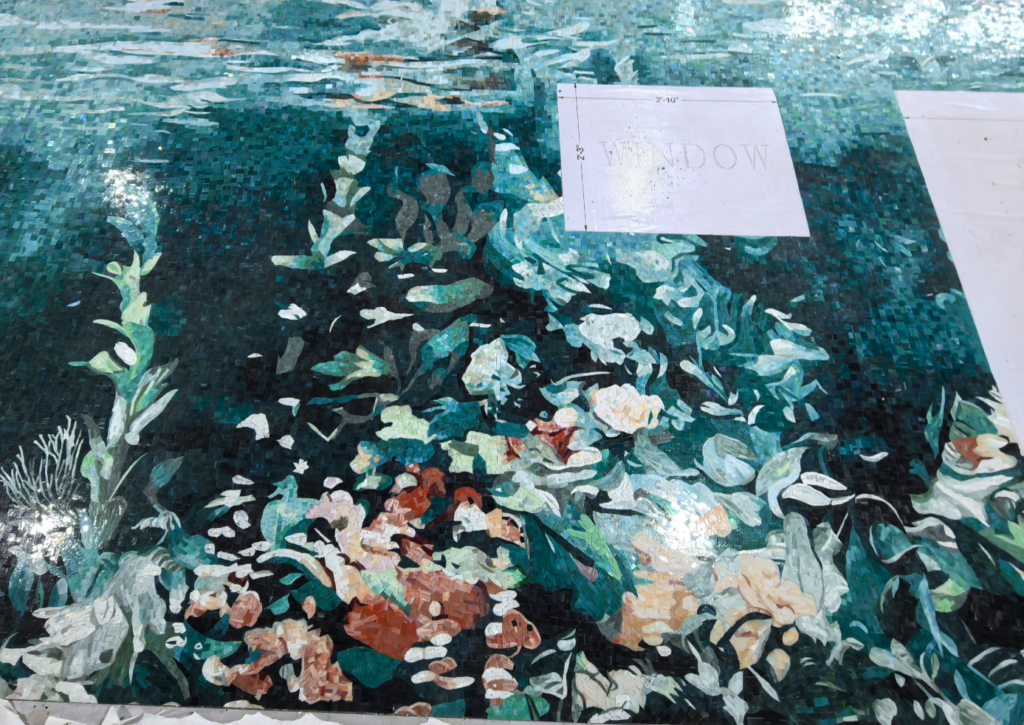 A close-up view of an in-production glass mosaic artwork laying flat in a workshop, showcasing rich rust-orange, deep teal, and cream tiles with architectural window markers.