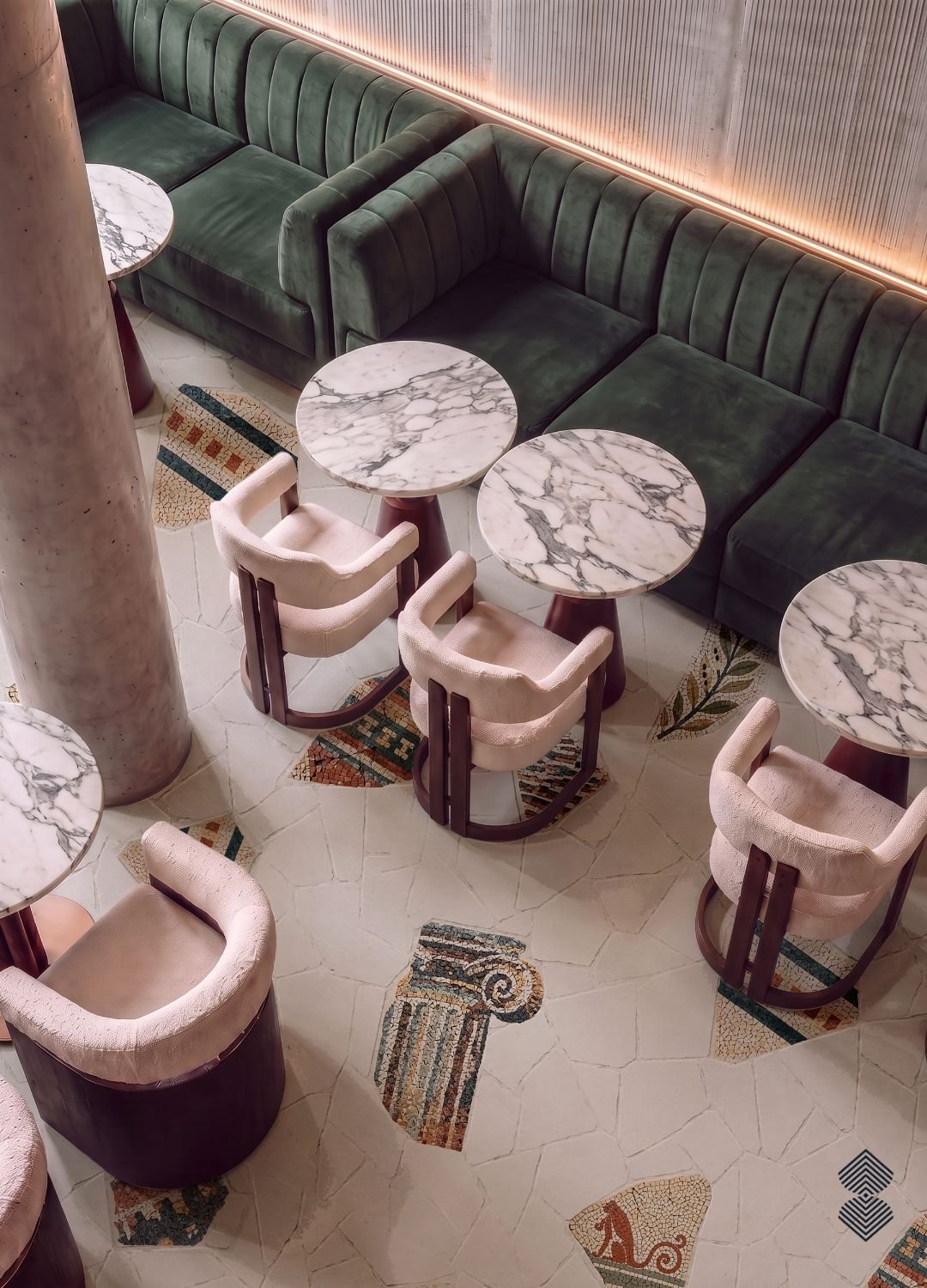 High-angle view of a chic restaurant lounge featuring plush green velvet tufted banquettes, round marble tables, and light pink armchairs over patterned mosaic tile flooring.