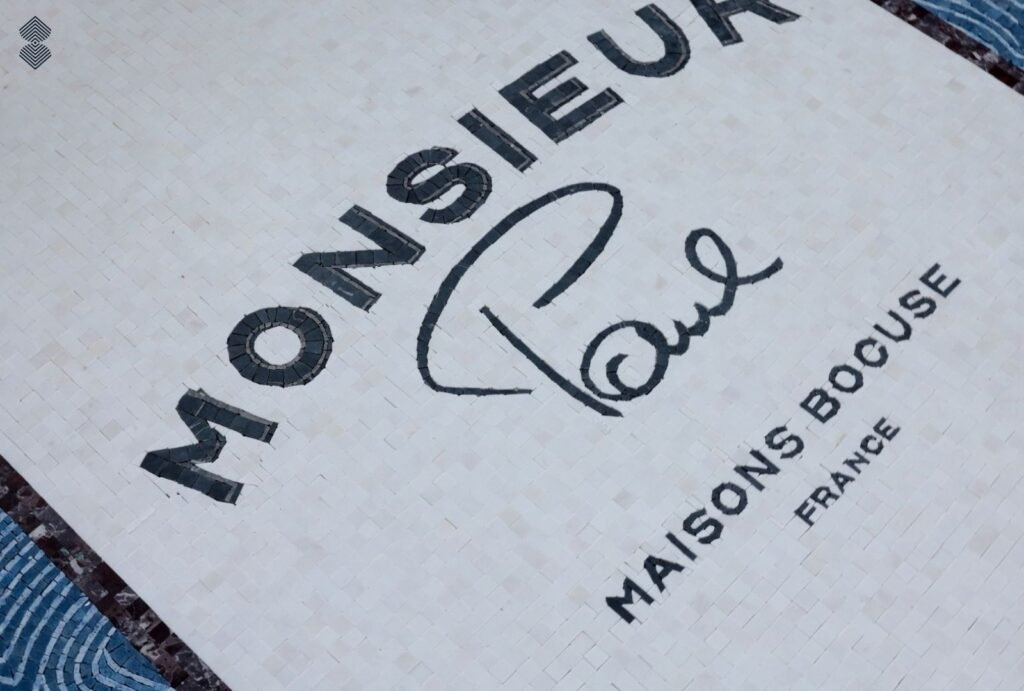 Close-up of light-colored mosaic floor tile inlaid with black text reading "MONSIEUR Paul" and "MAISONS BOCUSE FRANCE".