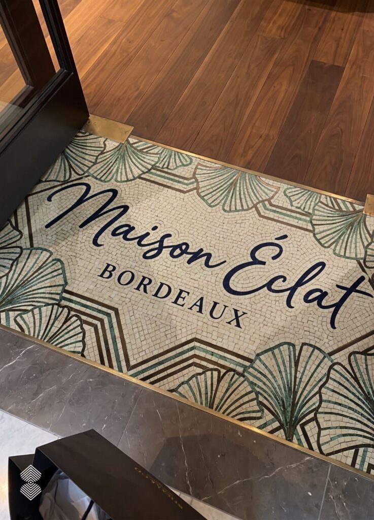 Floor inlay featuring Art Deco ginkgo leaf mosaic tile patterns surrounding black script text that reads "Maison Éclat BORDEAUX".