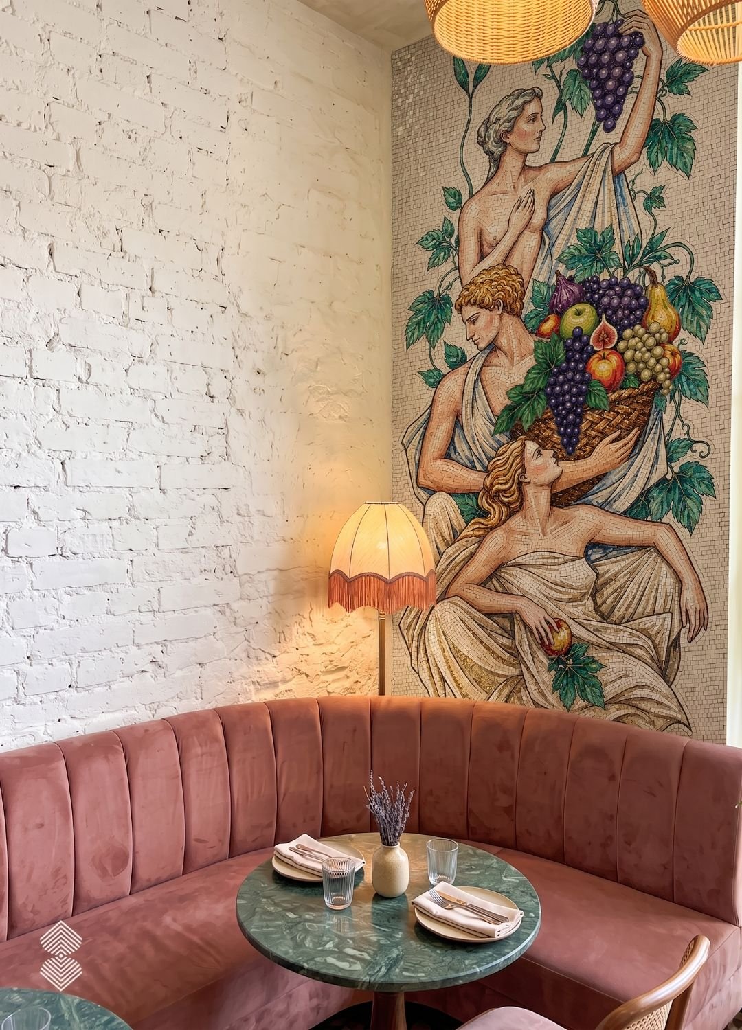 Corner booth with a pink channel-tufted banquette and a green marble table set against a classical mosaic mural of figures carrying fruit.