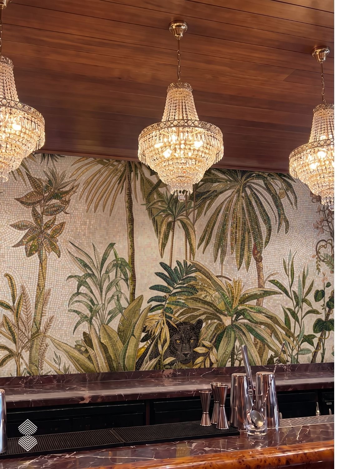 A bar counter illuminated by warm crystal chandeliers, backed by a detailed mosaic mural featuring tropical palm trees and a black panther.